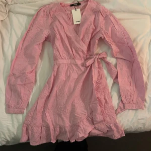 Rosa omlottklänning DW Riviera - Supersöt rosa omlottklänning från DW Riviera med knyt i midjan och volangkant nertill. Klänningen har långa ärmar och broderade blommor i tyget. Perfekt för vår och sommar!