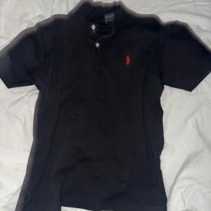 Svart pikétröja från Ralph Lauren - Klassisk svart pikétröja från Ralph Lauren med korta ärmar och röd broderad logga på bröstet. Tröjan har krage och två knappar framtill. Perfekt för en stilren och sportig look.