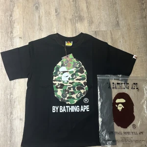 Svart t-shirt från BAPE med camo-tryck - Säljer en svart t-shirt från BAPE med stort kamouflage-tryck och klassisk BAPE-logga på bröstet. T-shirten har rund hals och korta ärmar. Märkeslapp på ärmen och tryckt text 'BY BATHING APE' nedtill.