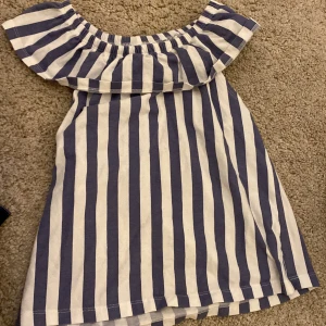 Randig klänning från Lindex Kids - Superfin klänning från Lindex Kids i vitt och blått med breda vertikala ränder. Klänningen har en volang upptill och är axelbandslös, perfekt för varma dagar. Mjuk och skön bomullskvalitet.