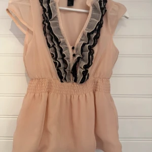 Beige blus från H&M - Säljer en söt beige blus från H&M med volangdetaljer i svart och vitt längs den v-ringade halsen. Blusen har korta ärmar och en elastisk midja som ger en fin passform. Inga defekte!!💞💞