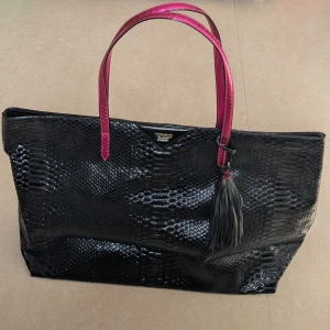 Shoppingväska Victoria's Secret  - Stor svart shoppingväska med glansig ormskinnsimitation och rosa handtag. Väskan har tofsdetalj och flera innerfack för enkel organisering. Perfekt för dig som vill ha plats med mycket och samtidigt sticka ut med färgglada detaljer. Väskan har aldrig använts 
