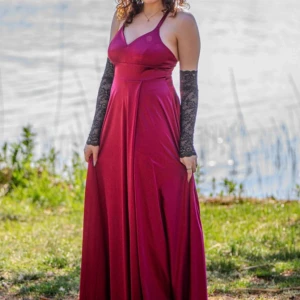 Vinröd långklänning med v-ringning - Säljer en vinröd långklänning med v-ringad hals och öppen rygg. Klänningen har smala axelband som korsas i ryggen och en hög slits framtill. Materialet är glansigt och faller snyggt. Perfekt för bal eller fest.