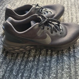 Svarta Nike sneakers med snörning - Säljer ett par svarta Nike sneakers med rund tå och platt sula. Skorna har en diskret Nike-logga på sidan och är tillverkade i syntetmaterial. Perfekta för träning eller vardagsbruk.