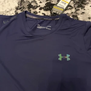 Blå t-shirt från Under Armour - Säljer en blå t-shirt från Under Armour med logga på bröstet. T-shirten har rund halsringning och är tillverkad i ett lätt och syntetiskt material som passar perfekt för träning.