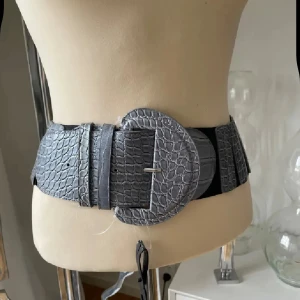 Grått bälte med krokodilpräglat mönster och stor rund spänne - Säljer ett brett grått bälte med krokodilpräglat mönster och stor rund spänne. Bältet har elastisk baksida för bekväm passform och är perfekt för att ge extra edge till din outfit.