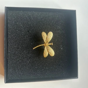 Guldfärgad ring i form av trollslända - Säljer en snygg guldfärgad ring formad som en trollslända. Ringen har detaljerade vingar med struktur och fäste på baksidan. Perfekt accessoar för att ge din outfit en unik touch.