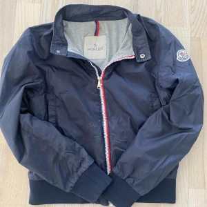 Mörkblå vindjacka från Moncler - Snygg mörkblå vindjacka från Moncler med klassisk logotyp på ärmen och dragkedja framtill med röd, vit och blå detalj. Jackan har ribbade muddar och krage, samt två sidofickor. Perfekt för dig som vill ha en stilren och sportig look. Ny pris 3500kr