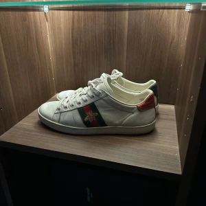 Gucci Ace sneakers - Gucci Ace sneakers i vitt skinn med gröna och röda detaljer på hälen. Skorna är i gott skick och är 7/10 skick