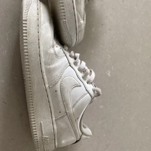 Vita Nike Air Force 1 sneakers - Klassiska vita Nike Air Force 1 sneakers med låg profil och rund tå. Skorna har snörning och är tillverkade i skinn med en platt sula. Perfekta för en stilren och sportig look.