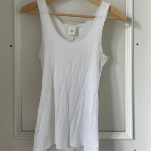Vit linnetopp med spetskant från H&M - Säljer en klassisk vit linnetopp från H&M L.O.G.G. med fina spetsdetaljer längs ringningen och axelbanden. Perfekt att styla till olika outfits under sommaren. Mjuk och skön bomullskvalitet.