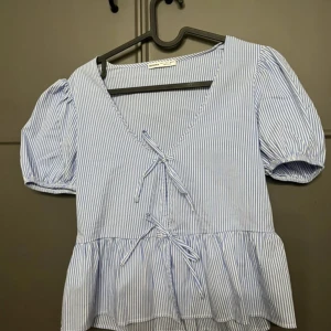 Blårandig blus med knyt och volang - Söt blus med blåvita ränder, puffärmar och två knyt framtill. Djup v-ringning och volang nertill ger en romantisk känsla. Perfekt för vår och sommar!