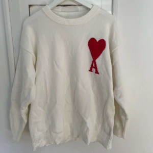 Vit stickad tröja med rött hjärta från Ami Paris - Vit stickad tröja från Ami Paris med ett stort rött hjärta och bokstaven A broderat på bröstet. Perfekt för en stilren och trendig look.