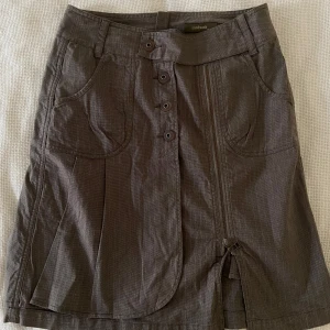 Brun kjol med knappar från Sandwich - Snygg grå kjol från Sandwich med dekorativa knappar framtill och dragkedja. Kjolen har två fickor fram och en stilren design som passar perfekt till vardags. Materialet känns lätt och bekvämt.