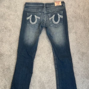 Blå jeansbyxor från True Religion - Säljer ett par klassiska blå jeans från True Religion med tydliga kontrastsömmar och deras ikoniska broderade bakfickor. Jeansen har låg midja och bootcut-stil. Säljer då dem inte passar och använt 3-5 gånger. Storlek 28 men dem sitter lite tight så  skulle säga att dem sitter som en 27a