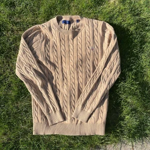 Beige kabelstickad tröja från GANT - Säljer en klassisk beige kabelstickad tröja från GANT. Tröjan har rund halsringning, ribbade muddar och ett litet broderat GANT-emblem på bröstet. Perfekt för lager-på-lager och snygg till jeans eller kjol.