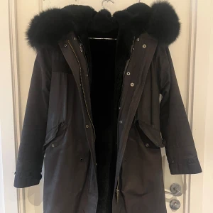 Svart parkajacka med pälsluva - Säljer en svart parkajacka med stor, fluffig pälskantad huva och dragkedja framtill. Jackan har långa ärmar, fickor med lock och knappar samt justerbar midja. Perfekt för kalla dagar. I storlek S 🩷