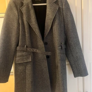 Grå kappa från Massimo Dutti med bälte - Säljer en klassisk grå kappa från Massimo Dutti med bälte i midjan och snyggt mönster. Kappan har lång ärm, krage och knappar framtill. Perfekt för dig som gillar stilrena och tidlösa plagg.