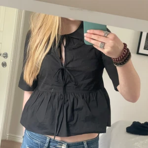 Svart blus med knytning - Svart blus med kort ärm och knytning framtill. Blusen har en lös passform och volang nedtill, samt en liten krage. Perfekt till jeans eller kjol för en trendig look.