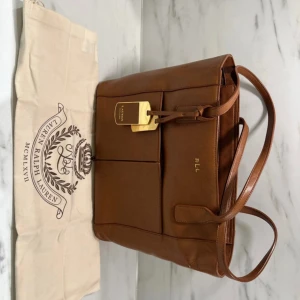 Brun handväska från Lauren Ralph Lauren - Snygg brun handväska i skinn från Lauren Ralph Lauren med guldfärgade detaljer och logotyp. Väskan har dubbla handtag, flera innerfack med dragkedja och en klassisk, stilren design. Perfekt accessoar för att lyfta din outfit.