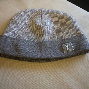 Grå rutig mössa från Louis Vuitton - Snygg grå mössa från Louis Vuitton med diskret rutigt mönster och uppvikt kant. LV-logga framtill i silver. Perfekt accessoar för kyliga dagar.