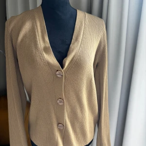 Vringad kofta - Säljer en beige ribbad kofta från Gerard Darel med v-ringning och tre knappar framtill. Koftan har lång ärm och är tillverkad i ett mjukt, stretchigt material som sitter snyggt på kroppen. Super trendig, perfekt till våren