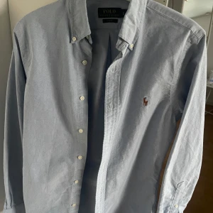 Ljusblå skjorta från Polo Ralph Lauren - Klassisk ljusblå skjorta från Polo Ralph Lauren i slim fit. Skjortan har knappar framtill, lång ärm och den ikoniska broderade loggan på bröstet. Tillverkad i bomull och passar perfekt till en clean och stilren look.