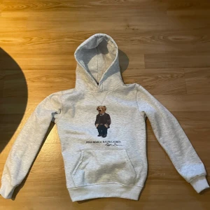 Grå hoodie Polo Bear Ralph Lauren S - Säljer en ljusgrå hoodie från Polo Ralph Lauren i storlek S, har du några frågor är det bara att fråga. Tröjan har den ikoniska Polo Bear-trycket på bröstet, känguruficka och huva. Mycket bra sick på tröjan 