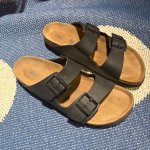 Svarta Birkenstock sandaler strl 43 - Klassiska svarta Birkenstock sandaler med två breda remmar och justerbara spännen. Ovandel i skinn och innersula i mocka för extra komfort. Sula i svart gummi med mönstrad undersida. Perfekta för sommaren och chill dagar.