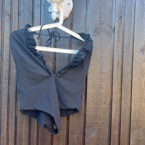 Svart baddräkt med volangdetalj - Supersnygg svart baddräkt med djup v-ringning och volangdetaljer längs kanten. Modellen har öppen rygg och knytband i nacken för en extra cool look. Perfekt för stranden eller poolen i sommar!