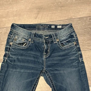 Miss Me jeans med broderad bakficka - Snygga blå jeans från Miss Me med kontrastsömmar och broderad bakficka med pärldetaljer. Jeansen har låg midja, fransade benslut och klassisk femficksdesign. Perfekta för dig som vill sticka ut med coola detaljer.