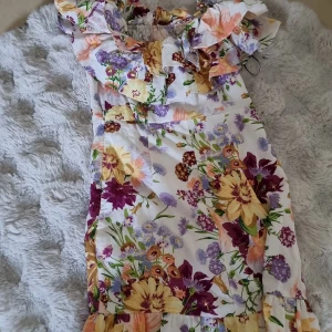 Blommig kortklänning från H&M - Superfin kortklänning från H&M i storlek 34 med färgglatt blommönster i lila, gul, orange och vinrött på vit botten. Klänningen har volang vid axlarna som är stretching. Perfekt för sommaren. Använt en gång.