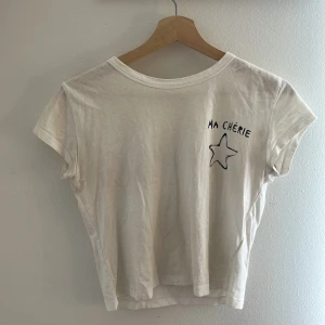 Vit t-shirt Ma Chérie Gina Tricot - Vit t-shirt från Gina Tricot med svart tryck 'MA CHÉRIE' och en stjärna både fram och bak. Modellen är kortärmad och har en klassisk rund halsringning. T-shirten är i mjuk bomull och har en avslappnad passform.
