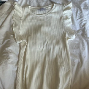 Vit ribbad topp med volangärmar Holly & Whyte - Supersöt vit ribbad topp från Holly & Whyte med volangdetaljer på axlarna. Toppen är ärmlös och har en rund halsringning. Perfekt för dig som gillar en feminin och stilren look med extra detaljer. Aldrig använd har endast tagit bort prislappen tyvärr.