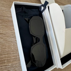Svarta solglasögon från Prada - Snygga svarta solglasögon från Prada med rektangulära, breda bågar och diskret logga på sidorna. Kommer med originalfodral och box. Perfekta för dig som vill ha en stilren och lyxig accessoar till din look.