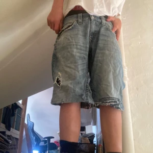 True religon shorts - Baggy blå jeansshorts med slitningar och hål på ena benet. Råa, fransiga kanter och klassiska femficksdetaljer. Perfekt för dig som gillar en avslappnad och streetig stil. Längre modell som går ner till knäna. Har sytt dom lite baggy men det syns knappast coola slytningar 
