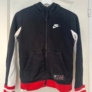 Nike Air svart hoodiejacka  - Svart hoodiejacka från Nike Air med vita och röda detaljer. Jackan har dragkedja framtill, huva, vita ärmar med röda ränder vid mudden och fickor framtill. Stor Nike Air-tryck på ryggen och liten logga på bröstet. Perfekt för en sportig och chill stil.