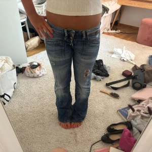 Lågmidjade bootcut jeans blå - Säljer ett par blå lågmidjade jeans med bootcut-snitt och slitna detaljer på benen. Jeansen har dubbla knappar i midjan och klassiska fickor fram och bak. Perfekta för dig som gillar en avslappnad och trendig look.