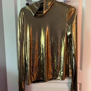Guldig glansig polotröja - Säljer en riktigt cool guldig polotröja med glansig finish. Tröjan har lång ärm och hög krage, perfekt för dig som vill sticka ut och ge din outfit en futuristisk touch. Materialet är stretchigt och följsamt, vilket gör den bekväm att bära.