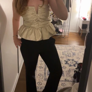 Guldig axelbandslös peplum tubtopp - Supersnygg axelbandslös tubtopp i guldigt, glittrigt tyg med struktur. Toppen har en markerad v-ringning, rynkade detaljer framtill och en volangkant nedtill som ger peplum-effekt. Perfekt för att sticka ut och skapa en glam look.