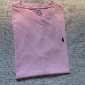 Rosa t-shirt från Polo Ralph Lauren - Snygg ljusrosa t-shirt från Polo Ralph Lauren med klassisk rund hals och ikoniska broderade loggan i mörkblått på bröstet. Tillverkad i mjuk bomull, perfekt för en clean och stilren look.