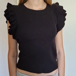 Svart topp med volangärmar - Svart topp med rund hals och korta volangärmar som ger en cool och trendig look. Toppens material är mjukt och stretchigt, ribbat, perfekt för att lyfta en enkel outfit. 