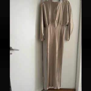 långklänning i satin - Elegant beige långklänning med långärmad design och rund halsringning. Klänningen har en snygg, figurnära passform och är tillverkad i glansigt satinmaterial som ger en lyxig känsla. Dragkedja baktill för enkel påklädning. Ny med prislapp. Klänningen är liten i storleken.