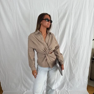 Beige skjorta med knytdetalj - Stilren beige skjorta i bomull med lång ärm och snygg knytdetalj framtill som ger en cool twist. Skjortan har klassisk krage och en loose fit, perfekt att styla med jeans eller kjol för en trendig look.