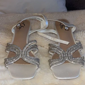 Glittriga vit silvriga klackar - Snygga vita klackar från din sko med fyrkantig tå och glittriga remmar täckta av små silvriga stenar. Skorna har en låg klack på ca 5 cm och justerbar rem runt ankeln med dekorativt spänne. Perfekta för att sticka ut med extra bling. Aldrig använda och skickar dirket efter köp