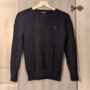 Intressekoll - SKRIV TILL MIG VID INTRESSE. Kabelstickad mörkblå tröja från Polo Ralph Lauren med klassisk rund hals och röd broderad logga på bröstet. Tröjan har ribbade muddar och är långärmad, perfekt för lager-på-lager under kyliga dagar.