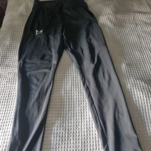 Byxor från Under Armour - Byxor från Under Armour med dragsko i midjan och vita logga på låret. Byxorna har smal passform, sidofickor och vita detaljer längs benen.