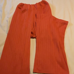 Roströda ribbade byxor Laura Kent - Säljer ett par roströda ribbade byxor från Laura Kent i storlek 36. Byxorna har bred resår i midjan och vida ben för en chill och trendig look. Perfekta för dig som gillar bekväma och snygga plagg med lite extra attityd.