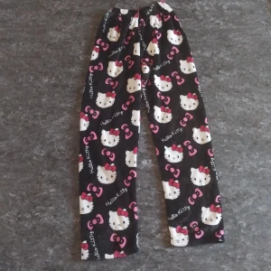 Hello Kitty fleecebyxor svart/rosa - Mysiga svarta fleecebyxor med Hello Kitty-motiv och rosa rosetter över hela byxan. Byxorna har resår i midjan och är perfekta för chill dagar hemma. Materialet är supermjukt och värmande, med ett gulligt och lekfullt mönster.