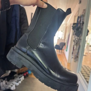 Svarta chunky hm boots i skinn - Säljer ett par svarta hm boots med chunky sula och elastiska paneler på sidorna. Skorna är i skinn och har en rund tå samt dragflikar både fram och bak för enkel påtagning. 